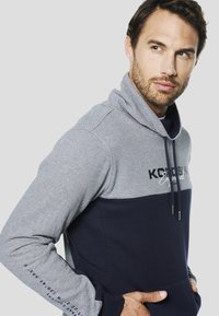 Sudadera con capucha con sección superior a rayas en tonos de gris y azul marino, con un bolsillo delantero y detalle de logo en el pecho.