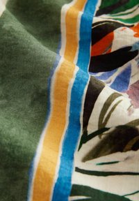 Gros plan sur un tissu avec une base verte et un motif rayé coloré en bleu, jaune, noir, blanc et orange accompagné de formes abstraites.