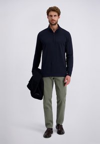 Marineblauwe longsleeve quarter-zip top, gecombineerd met olijfgroene broeken. Model houdt een zwarte jas vast. Glad stof, minimalistisch ontwerp en relaxte pasvorm.