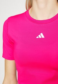 Jasně růžové sportovní tričko nošené osobou, se středovým bílým logem Adidas poblíž límce.