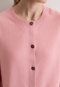 Cardigan rose clair en matériau doux, avec un col côtelé, cinq boutons marron foncé et un motif en tissu texturé.