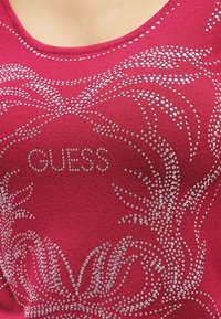 Top fucsia con scollatura rotonda, decorato con strass argento in un motivo floreale e il logo "GUESS" al centro. Tessuto testurizzato e elastico.