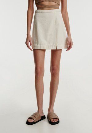 Vrouw draagt een beige minirok met een splitsing aan de voorkant en bruine gevoerde slidesandalen, staande tegen een effen lichte achtergrond.