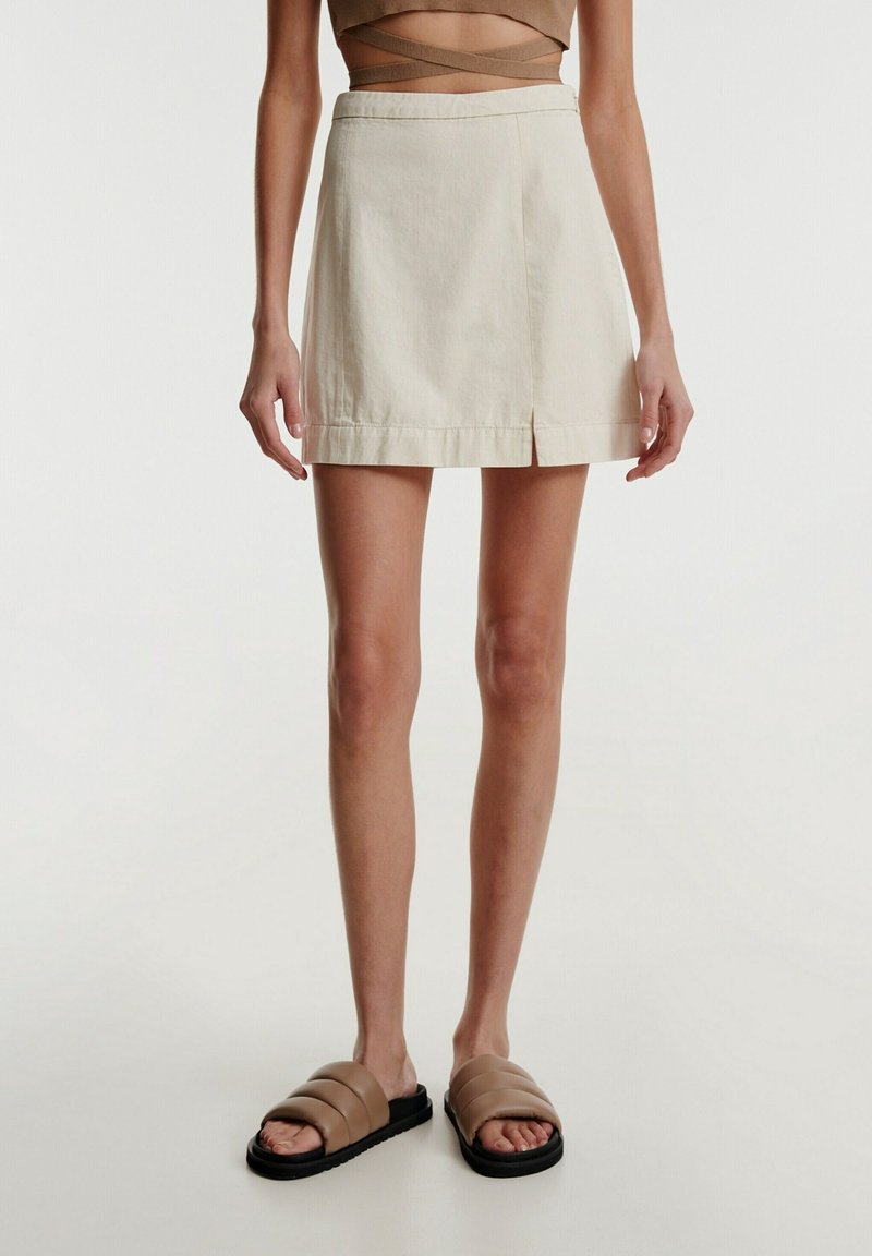 Femme portant une mini-jupe beige avec une fente à l’avant et des sandales slides marron matelassées, debout devant un fond clair uni.
