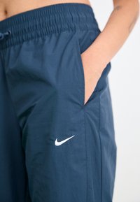 Tamsiai mėlynos Nike kelnes su elastinga juosmele ir šonine kišene, su baltu swoosh logotipu. Pagamintos iš lengvos, lygaus audinio.