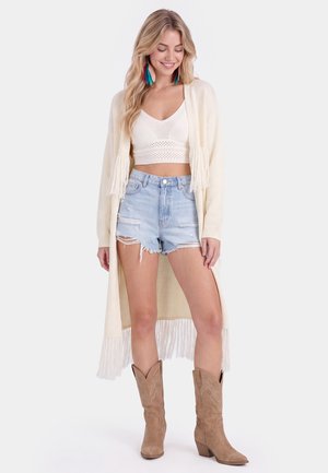 Smilende kvinde iført en hvid cropped top, lang cremefarvet cardigan med frynser, revnede denimshorts, brune cowboystøvler og farverige øreringe med fjer.