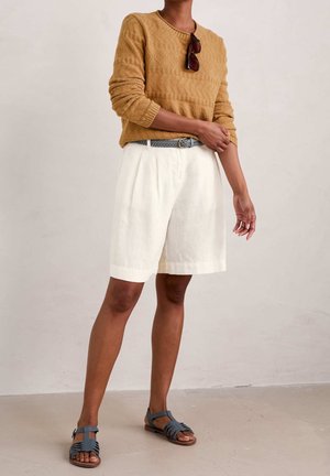 Persoon draagt een mosterdgeel trui met structuur, witte knielange shorts met een geweven riem, een zonnebril die aan de trui hangt, en blauwe sandalen.
