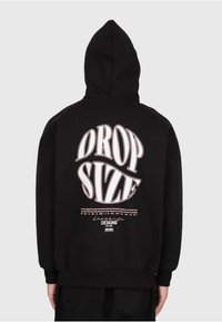 Persona che indossa un felpa nera con il cappuccio alzato, con testo grande bianco e rosa "DROP SIZE" e un design di testo più piccolo sulla schiena, su uno sfondo semplice.