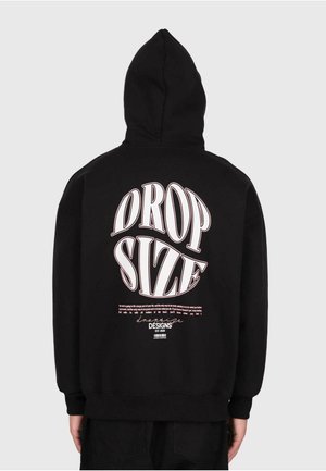 Persona che indossa un felpa nera con il cappuccio alzato, con testo grande bianco e rosa "DROP SIZE" e un design di testo più piccolo sulla schiena, su uno sfondo semplice.