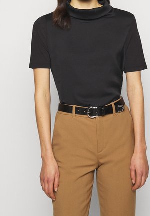 Persoon draagt een zwarte korte mouw coltrui, ingestopt in een hoog getailleerde beige broek met een zwarte riem met een zilveren gesp.