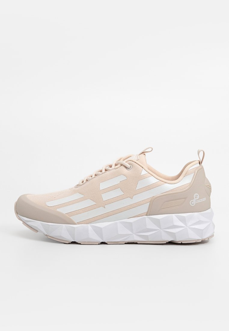 Zapatilla deportiva de color beige claro con parte superior de malla, acentos blancos, suela texturizada y patrón geométrico en la suela exterior. Diseño tradicional con cordones.