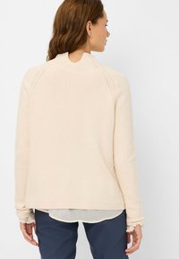 Maglione beige chiaro con colletto alto, bordi a coste e maniche lunghe. Presenta un tessuto lavorato a maglia e un look a strati con una camicia sottostante.