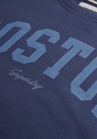 Sudadera azul marino con gran texto en azul claro que dice "OST" y un script más pequeño que dice "Superdry" debajo. Textura suave, cuello redondo.