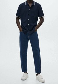 Camicetta a maniche corte blu navy con inserti bianchi nel colletto, tasca frontale, abbinata a jeans blu a gamba dritta e sneaker bianche. Design semplice e casual.