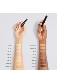 Dos brazos mostrando muestras de base, con múltiples tonalidades etiquetadas desde Clara hasta Oscura. Cada brazo sostiene un stick de base negro y rojo.