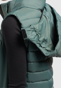 Kobieta w turkusowej kamizelce puchowej z dużym odpinanym kapturem, nosząca czarną bluzkę z długim rękawem, pokazana z boku od pasa w górę.