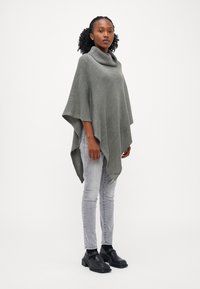 Grå strikket poncho med høy krage, med en løs passform og ujevn nederkant, kombinert med lysegrå smale jeans og svarte sko.