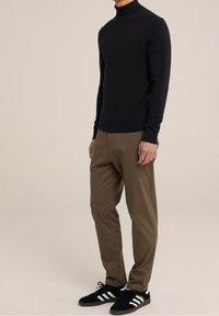 Man som bär en svart polo tröja, bruna byxor och svarta Adidas sneakers med vita ränder mot en enfärgad beige bakgrund.