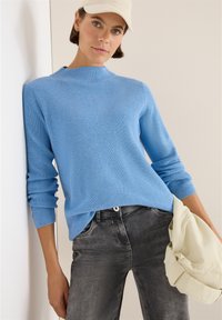 Blauer Strickpullover mit hochgeschlossenem Kragen, gerippter Textur und langen Ärmeln, kombiniert mit verwaschenen grauen Jeans und einer cremefarbenen Tasche.