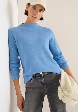 Blåstickad tröja med hög hals, ribbad textur och långa ärmar, kombinerad med blekta gråjeans och en krämfärgad väska.