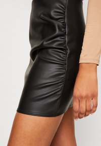 Mini jupe en simili cuir noir avec un détail de fronces sur le côté. Texture lisse, forme ajustée et couture visible à l'ourlet.