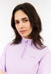 Donna con capelli scuri che indossa una felpa leggera viola con zip e il logo "protest" sul petto, guarda la macchina fotografica con un leggero sorriso.