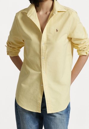 Person trägt ein hellgelbes Hemd mit aufgekrempelten Ärmeln und blaue Jeans, steht mit den Händen hinter dem Rücken.