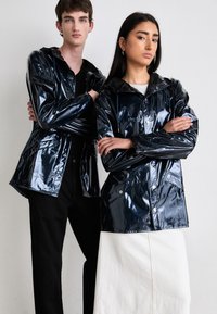 Glanzende zwarte, waterproof jassen met capuchons, voorzien van knopen aan de voorkant en zakken. Gedragen door twee modellen; de ene draagt het met zwarte broek, de andere met een witte rok.