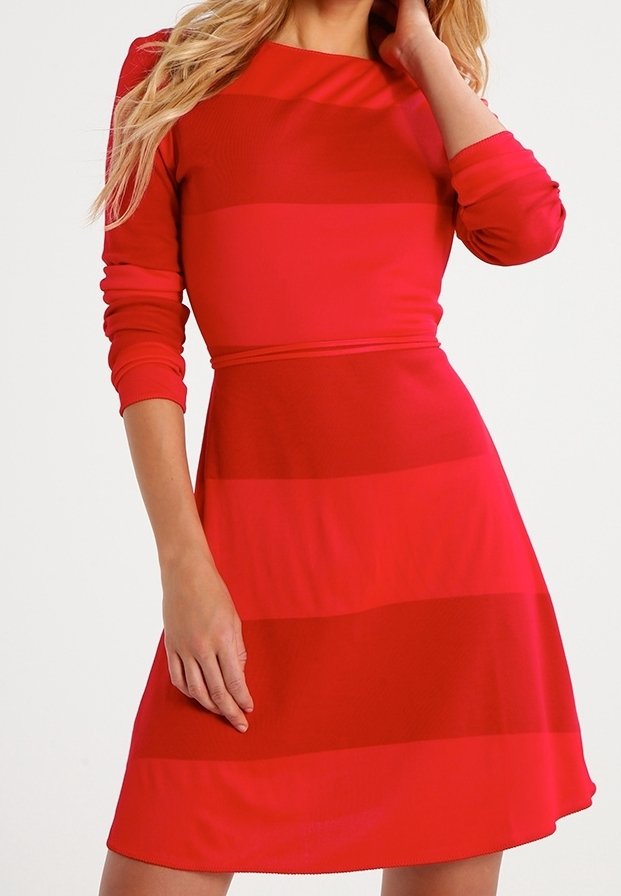 Femme portant une robe rouge à manches longues avec des rayures horizontales, une main reposant sur son cou et l’autre le long du corps.