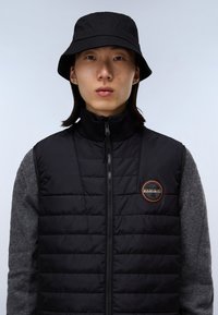 Svart quiltad väst med hög krage och dragkedja, tillsammans med en grå tröja. En rund logotyppatch syns på västen. Svart buckethat bärs.