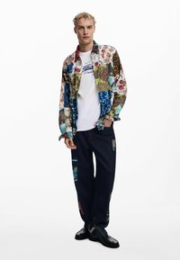 Camisa de patchwork colorido con patrones diversos, combinada con pantalones de mezclilla oscuros que presentan múltiples parches de tela y zapatos negros.