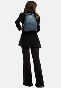 Blauwe leren rugzak met een gestructureerde afwerking, voorzien van twee schouderbanden en een voorste ritszak, gedragen over een zwarte outfit.