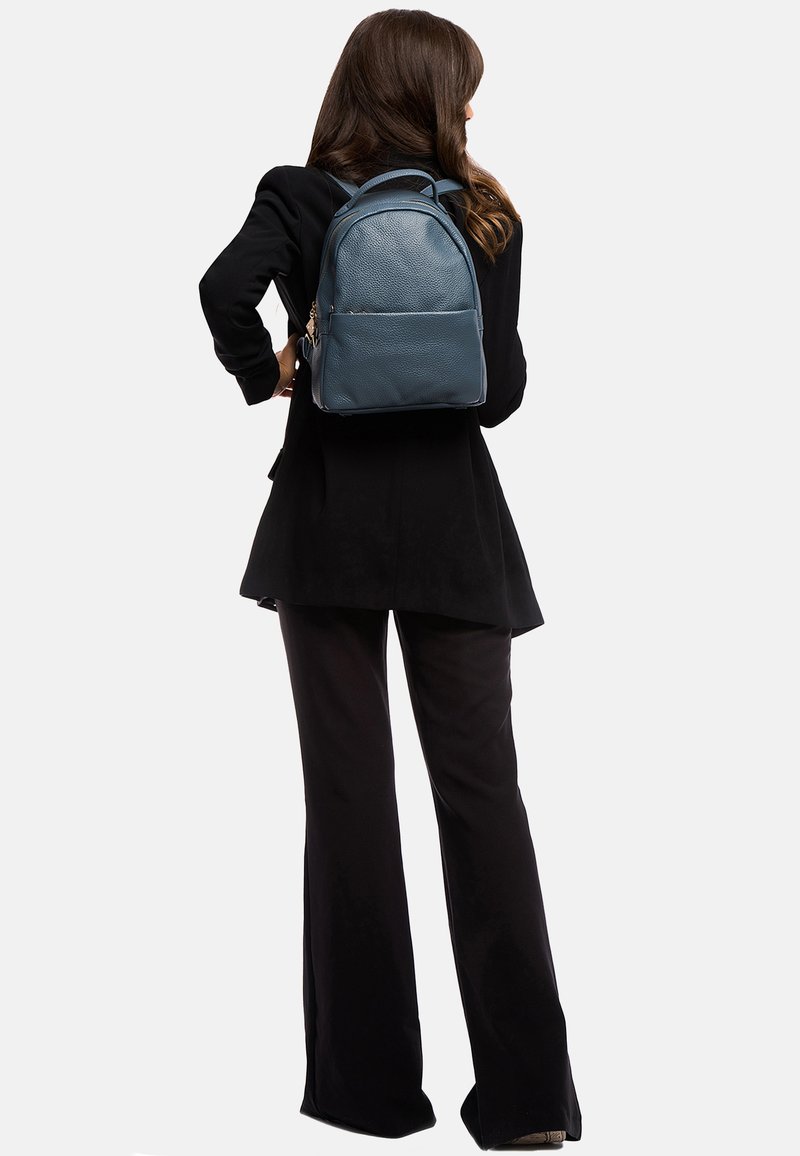 Blauwe leren rugzak met een gestructureerde afwerking, voorzien van twee schouderbanden en een voorste ritszak, gedragen over een zwarte outfit.