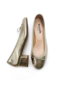 Zapatos planos metálicos dorados con un pequeño tacón cuadrado, punta redondeada y detalle de lazo. Textura suave con un forro interior suave. Logo de la marca en el interior.