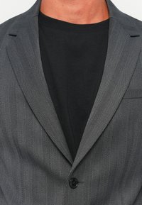 Grijs blazermodel met visgraatpatroon, voorzien van een zwarte knoop en notch revers, gelaagd over een zwart T-shirt met ronde hals. De stof is glad.