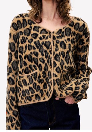 Mujer con un cárdigan de estampado de leopardo peludo con botones marrones y bolsillos delanteros, combinado con vaqueros azul oscuro, con la mano en el bolsillo.