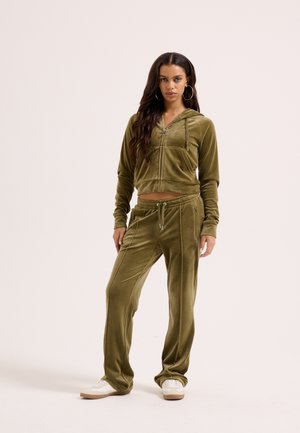 Žena stojaca oblečená v olivovo zelenej zamatovej mikine na zips a v zodpovedajúcich nohaviciach so šnúrkami, s bielymi teniskami na neutrálnom pozadí.