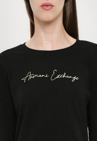 Black long-sleeve T-shirt featuring a round neckline and embroidered "Armani Exchange" logo in gold across the chest. Smooth cotton texture.