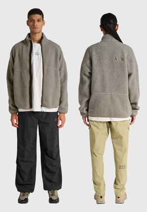 Man voor- en achterkant, gekleed in grijze fleecejassen met logo's, zwarte en beige broeken, en outdoor wandelschoenen tegen een effen achtergrond.