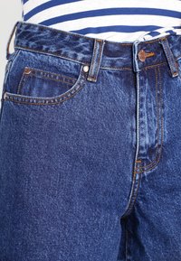 Vaqueros de mezclilla azul oscuro con un corte clásico, que cuentan con accesorios de cobre y un diseño de cinco bolsillos. Costuras visibles y fabricado de textura.