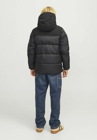 Črna jakna s kapuco in obloženimi rameni, s horizontalnim šivom, nosi jo skupaj z modrimi denim hlačami in rjavimi sandalmi s črnimi črtami.