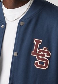 Chaqueta bomber azul marino con un aplique texturizado en burdeos y blanco con el logo "LS"; botones a presión plateados; llevada sobre una camiseta blanca.
