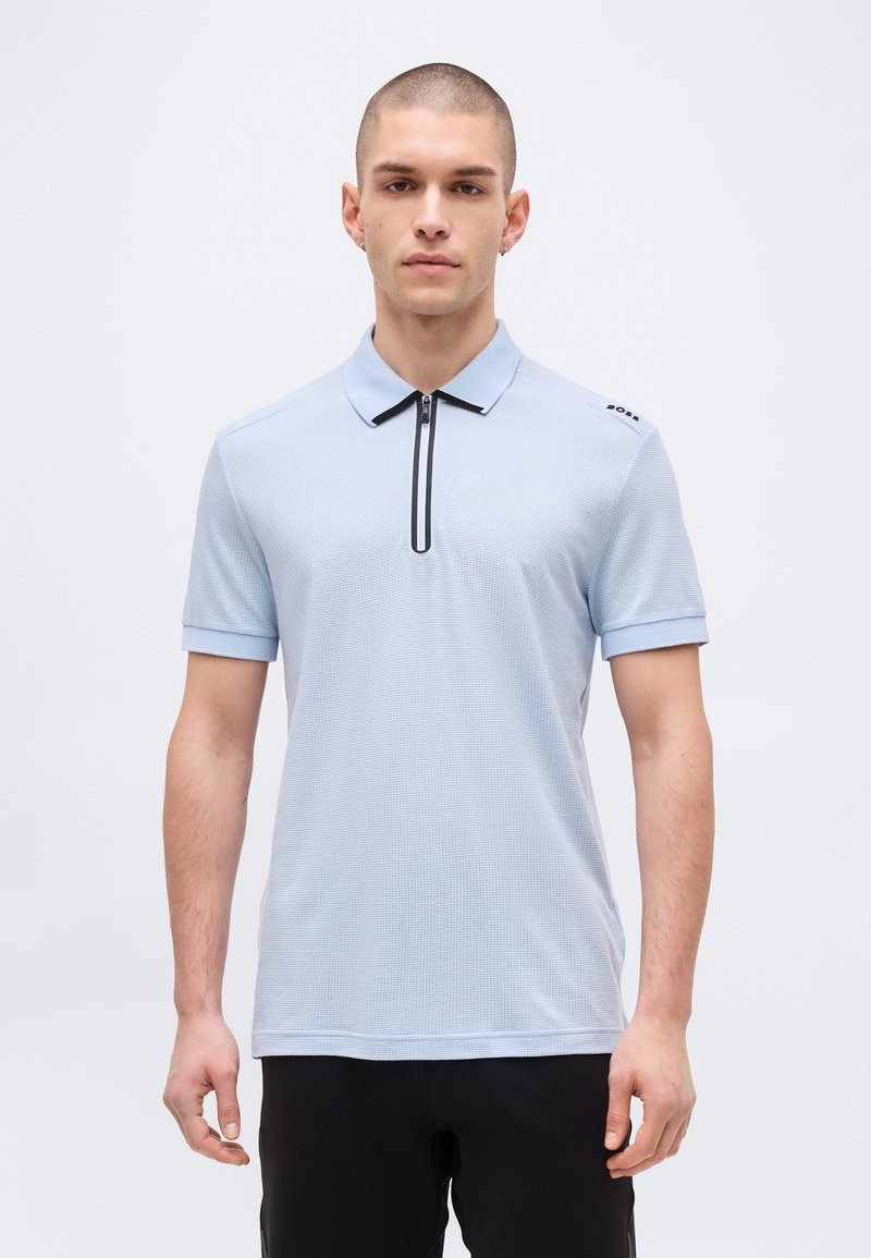 Homme portant un polo bleu clair à manches courtes avec un col zippé noir et un pantalon noir, debout devant un fond uni.