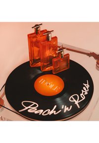 Tre bottiglie di profumo arancione di diverse dimensioni su un disco in vinile etichettato "IDÔLE" e testo scritto a mano "Peach'n Roses" su un giradischi.