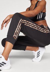 Černé sportovní kalhoty s béžovými a hnědými pruhy s leopardím vzorem. Mají zúžený střih, elastické manžety a jsou kombinovány s bílými teniskami.