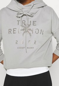 Felpa grigia con cappuccio in tessuto morbido, caratterizzata da decorazioni in strass. Il testo recita "TRUE RELIGION 2002 LUXURY BRAND" con dettaglio di palma.