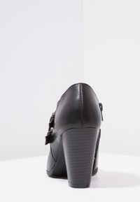 Zapato de tacón negro hecho de cuero sintético, con punta redondeada, tacón texturizado y detalles decorativos de hebilla.