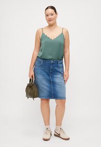 Zelena, valovita majica na bretele uparena s mid-length plavom traper suknjom. Mala maslinasta torba i bež tenisice upotpunjuju outfita.