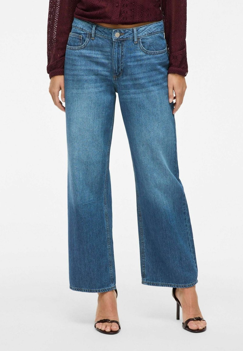 Femme portant un jean bleu taille mi-haute à jambes larges, un haut en maille bordeaux à manches longues et des sandales à talons noirs, debout devant un fond blanc.
