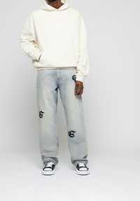 Uomo che indossa una felpa beige oversize, jeans chiari con toppe di lettere gotiche nere e sneaker bianco e nero, in piedi contro uno sfondo semplice.